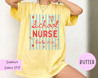 Puede incluir: Una camiseta de manga corta color amarillo mantequilla con un estampado gráfico de rayas verticales azul claro. El texto dice School Nurse, Emily y 2026 en rojo, junto con corazones rojos, un lazo rojo pequeño y un dibujo de un estetoscopio.
