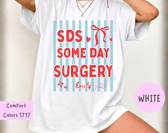 Puede incluir: Una camiseta blanca de manga corta con un fondo de rayas verticales azul claro y texto rojo que dice SDS SOME DAY SURGERY junto al nombre Emily en letra cursiva roja. El diseño incluye un lazo rojo, dos corazones pequeños, estrellas y un estetoscopio.