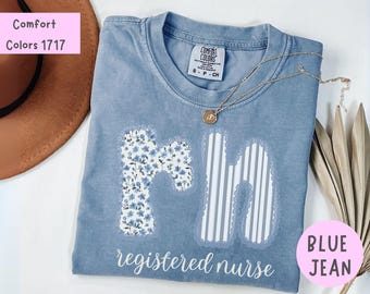 Puede incluir: Camiseta azul vaquero con la inscripción "registered nurse". Las letras "RN" están aplicadas con estampados florales y a rayas. Un collar dorado con un colgante descansa sobre la camiseta. La camiseta tiene una etiqueta Comfort Colors.