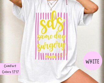 Puede incluir: Una camiseta blanca con un diseño gráfico de rayas verticales rosas y texto amarillo en cursiva que dice sds same day surgery y el nombre Paula. Un pequeño icono de estetoscopio amarillo se encuentra junto al nombre en la camiseta.