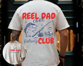 Puede incluir: Camiseta color crema con un diseño temático de pesca. El diseño incluye un pez azul, una caña de pescar, una caja de aparejos y las palabras "REEL DAD cool fishing CLUB" en rojo y azul. Una imagen más pequeña de la camiseta es visible en la esquina inferior izquierda.