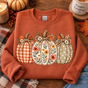 Puede incluir: Una sudadera de cuello redondo color óxido con un gráfico de tres calabazas. La calabaza izquierda tiene un patrón de cuadros vichy naranja, la central un estampado floral y la derecha un patrón de lunares verde salvia, atadas con cuerda.