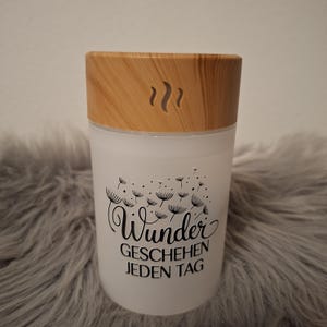 Op de afbeelding: Een witte cilindrische etherische olie diffuser met een houten deksel. De diffuser heeft een zwarte afbeelding van paardenbloemzaden en de woorden "Wunder GESCHEHEN JEDEN TAG". De deksel heeft een houtnerfpatroon en een stoom symbool.