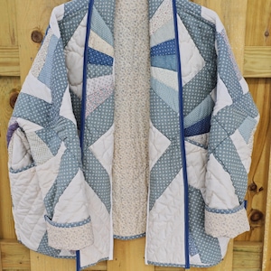 May include: A quilted jacket in shades of blue, white, and cream, with a geometric pattern. The jacket features a blue trim and cuffs, and is hanging on a wooden hanger. The interior lining is a cream color.