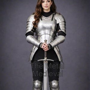 May include: A full suit of polished metal armor, including chest plate, shoulder guards, arm and leg protection. The armor is worn over a black top and pants. A long sword is held in front. The text "The Metal Medieval" is visible.