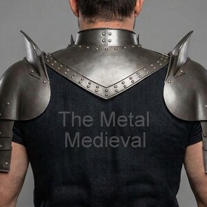 May include: A set of medieval-style metal shoulder armor, featuring a neck guard and articulated shoulder plates. The armor is a dark, metallic gray color, with rivets and pointed accents. The text "The Metal Medieval" is visible on the shirt.