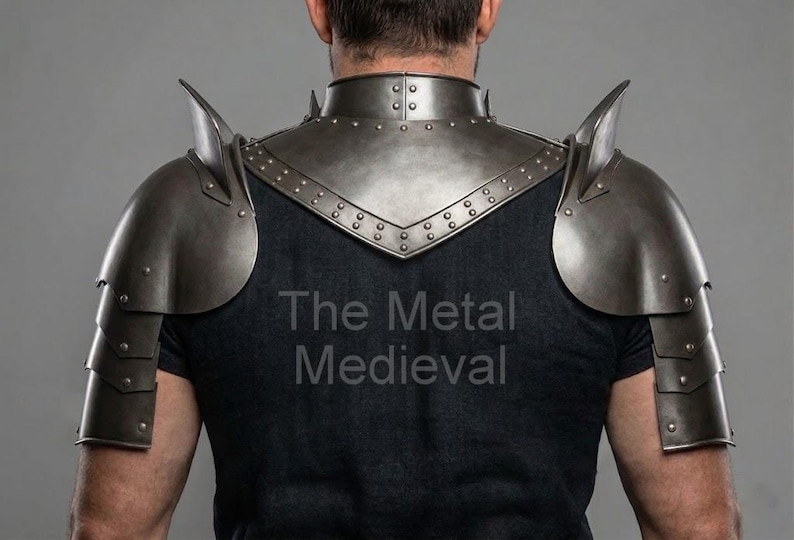 May include: A set of medieval-style metal shoulder armor, featuring a neck guard and articulated shoulder plates. The armor is a dark, metallic gray color, with rivets and pointed accents. The text "The Metal Medieval" is visible on the shirt.