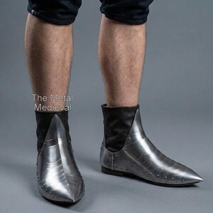 May include: A pair of polished metal medieval boots with a pointed toe and riveted construction. The boots have a black leather upper and are designed to protect the feet and ankles. The Metal Medieval logo is visible in the image.