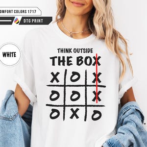 May include: White t-shirt featuring the phrase "THINK OUTSIDE THE BOX" above a tic-tac-toe grid. A red vertical line runs through the centre of the grid. The shirt also has the words "COMFORT COLORS 1717" and "DTG PRINT" printed on it.