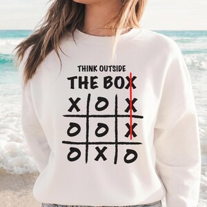 May include: White sweatshirt featuring the phrase "THINK OUTSIDE THE BOX" and a tic-tac-toe grid with X's and O's. A red line runs vertically through the grid. The sweatshirt is worn with blue jeans.