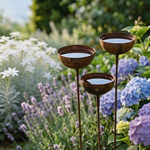 Op de afbeelding: Drie verweerde metalen vogelbaden met een ondiep komontwerp, gevuld met water. De vogelbaden staan op lange, slanke stelen, geplaatst in een weelderige tuin met lavendel, witte bloemen en blauwe en paarse hortensia's.
