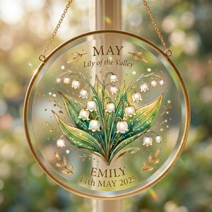 May include: A round, glass hanging decoration with a gold-colored metal frame and chain. The design features a Lily of the Valley bouquet with green leaves and white flowers. Text includes "MAY Lily of the Valley" and "EMILY 14th MAY 2025".