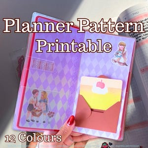 Puede incluir: Una funda de plástico transparente para agenda con un patrón de rombos morados y blancos, decorada con pegatinas de dos niños y un postre. Incluye un bolsillo de papel marrón con una tarjeta amarilla ilustrada con cereza y nata.