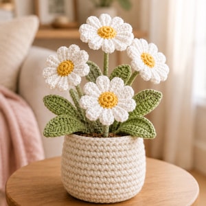 May include: A handmade crochet daisy arrangement in a cream-coloured pot. The daisies feature white petals and yellow centres. Green leaves and stems support the flowers. The pot sits on a wooden table, creating a charming, handmade decor piece.