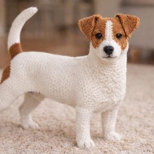 Op de afbeelding: Een gehaakte Jack Russell Terrier hond figuur. De hond is wit met bruine markeringen op zijn hoofd, oren en staart. Hij staat op een beige tapijt en heeft een geconcentreerde uitdrukking.