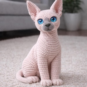 Puede incluir: Figura de gato esfinge hecha a mano con ganchillo, en hilo rosa claro. El gato tiene grandes ojos azules brillantes y una nariz gris. Está sentado sobre una alfombra blanca, con un fondo borroso que incluye un sofá y una planta.