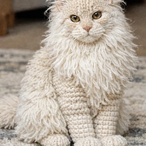 Può includere: Un gatto all'uncinetto fatto a mano, color crema, con dettagli. Il gatto ha una criniera soffice e un corpo strutturato, seduto in posizione eretta. Gli occhi del gatto sono color ambra e ha un piccolo naso rosa. Un oggetto decorativo.