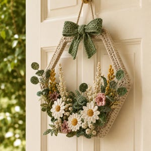 May include: A handmade, crochet floral wreath in a hexagonal shape. The wreath features white daisies, pink and yellow flowers, and green leaves. A green bow is attached at the top, and the wreath hangs from a beige cord.