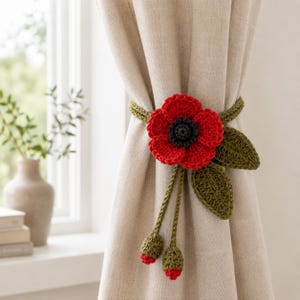 May include: A handmade, red crochet poppy flower curtain tieback with green leaves and buds. The flower has a black center and is attached to a green crocheted band. The tieback is designed to hold a curtain in place.