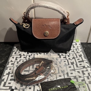 Könnte beinhalten: Schwarze Handtasche mit braunem Lederklappe und Griff. Die Tasche hat einen goldfarbenen Verschluss und einen abnehmbaren braunen Riemen. Die Tasche liegt auf einem schwarz-weiß gemusterten Papier.