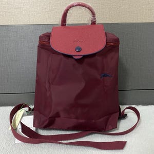 May include: A burgundy backpack with a red flap and handle. The flap is secured with a blue button. The bag has a small embroidered logo and adjustable straps. The handle is wrapped in clear plastic.