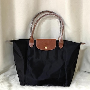 以下が含まれることがあります： 黒色のトートバッグ。茶色の革のアクセントがあり、金色のボタン留めが付いた茶色のフラップが付いています。茶色の革のハンドルとジッパー開閉部があります。耐久性のある素材で作られています。