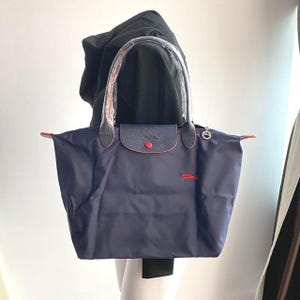 May include: Navy blue tote bag with red trim and a flap closure. The bag features a red logo and a silver-tone charm. The handles are clear. The bag is made of a durable material.