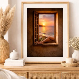 Puede incluir: Impresión artística enmarcada que muestra una vista del atardecer a través de una ventana abierta. La imagen muestra un cielo naranja y amarillo vibrante sobre el mar, con las piernas de una persona descansando en el alféizar. El marco es marrón claro.