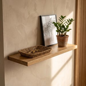 May include: A light brown wooden floating shelf holding a rectangular woven seagrass tray containing metal keys, a framed black and white line art print of a figure, and a green Zamioculcas plant in a terracotta pot against a textured wall.