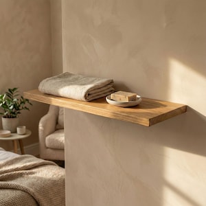 Könnte beinhalten: Ein Holzregal an einer beigefarbenen Wand, mit einem gefalteten beigefarbenen Handtuch und einer kleinen weißen Schale mit einem Stück Seife. Das Regal ist aus hellem Holz.