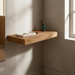 May include: A floating wooden shelf mounted on a light-colored wall. A white tray with keys, a small notebook, and a green jar with a pen are on the shelf. The shelf is made of natural wood and is installed in a corner.