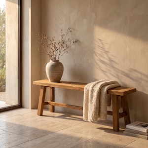 May include: A rectangular wooden bench with four sturdy legs and a horizontal support bar. A thick, cream-colored knitted throw blanket is draped over one end of the bench. A large, textured, light brown ceramic vase holds dried branches.