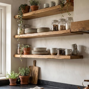 May include: Three natural wood floating shelves mounted on a wall, holding stacks of ceramic plates, glass storage jars with contents, various mugs, and potted green plants. A wooden cutting board rests on the counter below the bottom shelf.