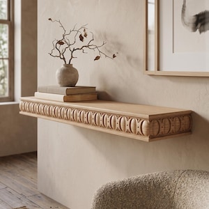May include: A light-colored wooden floating shelf featuring a decorative carved pattern along the front edge, consisting of repeating circular motifs. The shelf is mounted on a wall and holds two stacked books and a small textured vase with branches.
