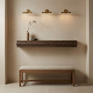 Op de afbeelding: Een kamerinterieur met een houten bank en een gebeeldhouwd houten plank. Boven de plank bevinden zich drie messing wandkandelaars. Een vaas en een kleine kom staan op de plank. De muur is geschilderd in een neutrale tint. De bank is 150 cm lang.