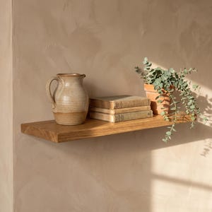 May include: A natural wood floating shelf holding a ceramic pitcher with a speckled beige finish, a stack of two hardcover books with aged pages, and a potted eucalyptus plant in a terracotta pot, all arranged on a textured beige wall.