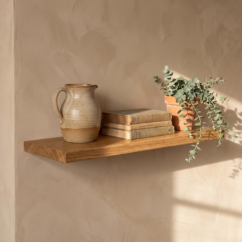 May include: A natural wood floating shelf holding a ceramic pitcher with a speckled beige finish, a stack of two hardcover books with aged pages, and a potted eucalyptus plant in a terracotta pot, all arranged on a textured beige wall.
