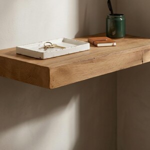 May include: A rustic wooden shelf with a white rectangular tray holding keys. A small brown notebook and a green ceramic jar with a pen are also on the shelf. The shelf is mounted on a light-colored wall.