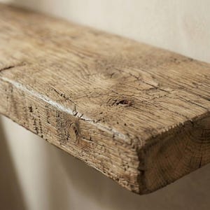 Pu&ograve; includere: Primo piano di una mensola in legno rustico. La mensola &egrave; realizzata in legno marrone chiaro, invecchiato, con venature, nodi e crepe visibili, che le conferiscono un aspetto naturale e invecchiato. La mensola &egrave; montata su una parete di colore neutro.