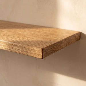 May include: A rectangular floating wall shelf made of natural light brown oak wood. The shelf features a visible wood grain pattern with a smooth, finished surface and a straight, clean edge, extending horizontally across the frame.