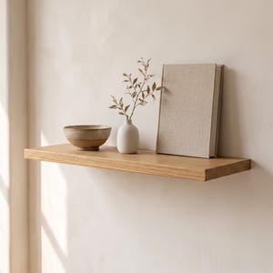 May include: A light-toned wooden floating shelf mounted on a wall. On the shelf sits a two-toned ceramic bowl with a dark base and light rim, a small white vase containing a delicate leafy branch, and a closed book with a textured beige cover.