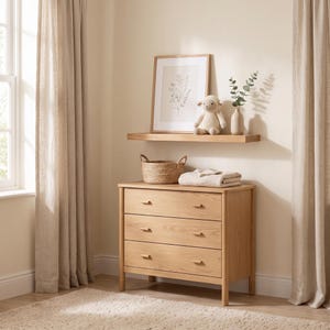 May include: A light oak wooden dresser with three drawers, topped with a woven basket, a folded beige blanket, and a wall-mounted shelf holding a framed botanical print, a plush cream-colored sheep toy, and a small vase with green leaves.