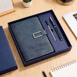 May include: A blue leather journal with a silver clasp, presented in a matching gift box. The journal is engraved with the text "Dr. Brown Ph.D. 2026". A silver pen and a black pen are included in the box.