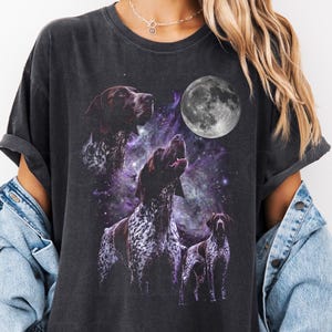 May include: A dark gray t-shirt featuring a graphic of German Shorthaired Pointers howling at the moon. The design includes a full moon and a purple galaxy background. The text "COLOR: PEPER" is on the left side.