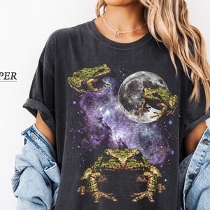 Può includere: Una maglietta grigio scuro con un design fantasy di rane sullo sfondo di una galassia e una luna. Le rane sono verdi e marroni, e la galassia è un mix di viola e blu. Il testo "COLOR: PEPER" è a sinistra.