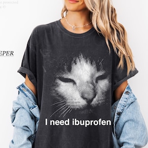 May include: A dark gray t-shirt with a distressed cat face graphic. The text "I need ibuprofen" is printed below the cat's face. The shirt is a pepper color.