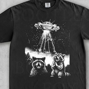 May include: Black t-shirt with a graphic of a UFO beaming light onto two raccoons. The illustration is in black and white. The shirt has a crew neck and short sleeves. The text "Washed Black" is at the bottom.