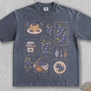May include: A charcoal gray short-sleeve t-shirt featuring a graphic print of various blueberry-themed illustrations, including a stack of pancakes, a jar of jam, a basket of berries, and sprigs of blueberries arranged in decorative rectangular frames.