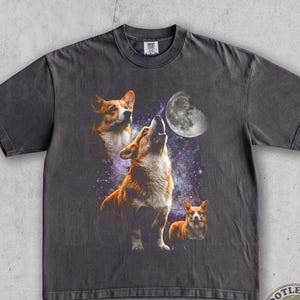 May include: A charcoal gray short-sleeve t-shirt featuring a graphic print of three Corgi dogs against a purple and black galaxy background with a large moon. The central Corgi is howling upward, while the others look toward the moon.
