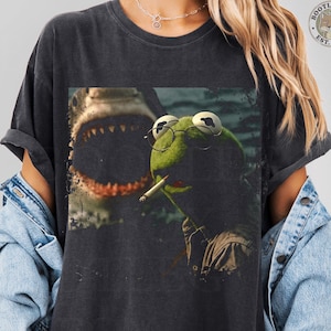 May include: Dark gray t-shirt featuring a humorous graphic of Kermit the Frog wearing glasses and smoking a cigarette, with a shark in the background. The shirt has a vintage, distressed look.